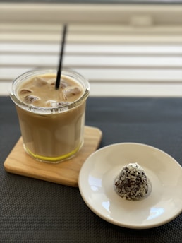 A glass of iced coffee with a straw sits on a wooden coaster, accompanied by a small chocolate-covered treat with coconut shavings on a white dish. The background features a striped pattern, creating a cozy and inviting atmosphere.