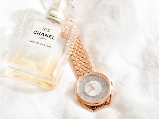 A bottle of Chanel No. 5 Eau de Parfum is placed next to a rose gold wristwatch with a textured face. Both items rest on a soft, white fabric background, emphasizing a sense of luxury and elegance.