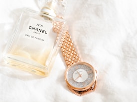 A bottle of Chanel No. 5 Eau de Parfum is placed next to a rose gold wristwatch with a textured face. Both items rest on a soft, white fabric background, emphasizing a sense of luxury and elegance.
