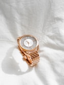 A gold wristwatch with a textured metallic face and a shiny bracelet strap rests on a crumpled white fabric background. The watch face is round and features delicate, subtle detailing.