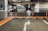 A well-lit underground parking garage with marked parking spaces. Several cars are parked near concrete pillars that are decorated with geometric patterns. Orange traffic cones and a directional arrow on the floor guide traffic flow. Overhead signage indicates limited clearance.