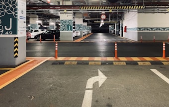 A well-lit underground parking garage with marked parking spaces. Several cars are parked near concrete pillars that are decorated with geometric patterns. Orange traffic cones and a directional arrow on the floor guide traffic flow. Overhead signage indicates limited clearance.