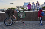 A person with a blue mohawk hairstyle is fixing a bicycle that is upside down on a paved walkway. Nearby, a group of children in warm clothing are observing the scene. The background includes a water fountain and modern buildings.