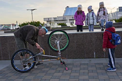 A person with a blue mohawk hairstyle is fixing a bicycle that is upside down on a paved walkway. Nearby, a group of children in warm clothing are observing the scene. The background includes a water fountain and modern buildings.