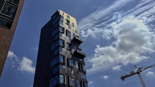 A tall, modern high-rise building with multiple windows and a unique façade stands against a partly cloudy sky. In the background, a construction crane is visible, indicating urban development or construction activity. The sky is blue with white, fluffy clouds and some wispy cloud formations.