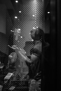 A person is standing in a recording studio. The individual wears headphones and is facing a professional microphone. The room is acoustically treated with foam padding on the walls. They appear to be gesturing with one hand in an expressive manner.