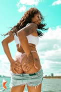 A woman with long curly hair and tattoos is by the water, wearing a white crop top and denim shorts. She has a confident and relaxed posture, with one hand on her waist. The background features a bright blue sky with some clouds and distant city buildings.