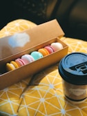 A box of assorted colorful macarons rests on a yellow geometric-patterned fabric surface. Next to the box is a takeaway coffee cup with a branded label, emphasizing a cozy, snack-time setting.