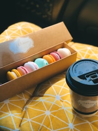 A box of assorted colorful macarons rests on a yellow geometric-patterned fabric surface. Next to the box is a takeaway coffee cup with a branded label, emphasizing a cozy, snack-time setting.