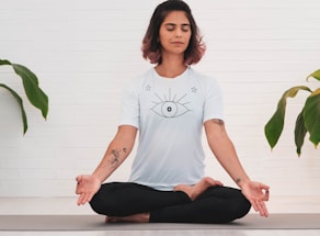 A person is meditating in a seated cross-legged position on a yoga mat. They have short, dark hair with reddish tips and are wearing a white t-shirt featuring a large eye design on the front. The person has tattoos on both arms. Surrounding them, there are green plant leaves against a white brick wall background.