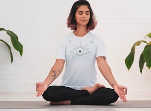 A person is meditating in a seated cross-legged position on a yoga mat. They have short, dark hair with reddish tips and are wearing a white t-shirt featuring a large eye design on the front. The person has tattoos on both arms. Surrounding them, there are green plant leaves against a white brick wall background.