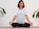 A person is meditating in a seated cross-legged position on a yoga mat. They have short, dark hair with reddish tips and are wearing a white t-shirt featuring a large eye design on the front. The person has tattoos on both arms. Surrounding them, there are green plant leaves against a white brick wall background.