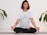 A person is meditating in a seated cross-legged position on a yoga mat. They have short, dark hair with reddish tips and are wearing a white t-shirt featuring a large eye design on the front. The person has tattoos on both arms. Surrounding them, there are green plant leaves against a white brick wall background.