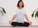 A person is meditating in a seated cross-legged position on a yoga mat. They have short, dark hair with reddish tips and are wearing a white t-shirt featuring a large eye design on the front. The person has tattoos on both arms. Surrounding them, there are green plant leaves against a white brick wall background.