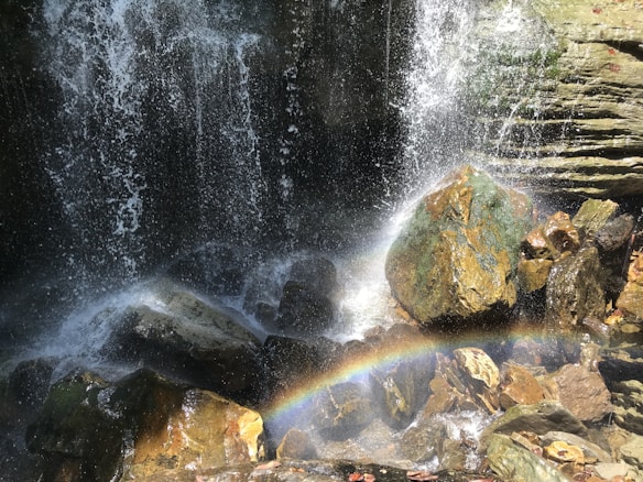 Water cascades down rocks, creating a mist that catches sunlight and forms a vivid rainbow. The scene is set against rugged, textured stones, some of which are mossy. The water splashes energetically, adding a sense of movement to the serene natural setting.