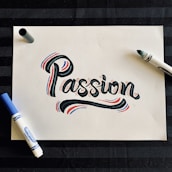 A piece of white paper features the word 'Passion' written in a bold, artistic style with black ink, accented by red and blue highlights. Surrounding the word are two markers, one blue and one black, situated on opposite sides of the paper. The background is a dark, striped surface.