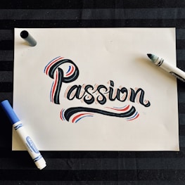 A piece of white paper features the word 'Passion' written in a bold, artistic style with black ink, accented by red and blue highlights. Surrounding the word are two markers, one blue and one black, situated on opposite sides of the paper. The background is a dark, striped surface.