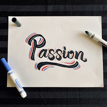 A piece of white paper features the word 'Passion' written in a bold, artistic style with black ink, accented by red and blue highlights. Surrounding the word are two markers, one blue and one black, situated on opposite sides of the paper. The background is a dark, striped surface.