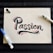 A piece of white paper features the word 'Passion' written in a bold, artistic style with black ink, accented by red and blue highlights. Surrounding the word are two markers, one blue and one black, situated on opposite sides of the paper. The background is a dark, striped surface.