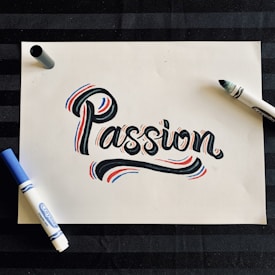 A piece of white paper features the word 'Passion' written in a bold, artistic style with black ink, accented by red and blue highlights. Surrounding the word are two markers, one blue and one black, situated on opposite sides of the paper. The background is a dark, striped surface.