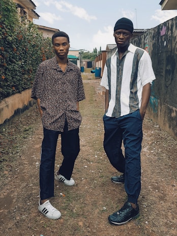 Two young men in athletic wear: one in a retro Argentina soccer jersey with Nike pants, the other in a vintage Boca Juniors jersey paired with retro Adidas pants, standing outdoors with urban street art behind.