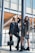Two stylishly dressed women pose confidently in front of a modern architectural backdrop. Both are wearing sunglasses and hold black designer handbags. One woman is wearing a black fur coat and leather pants, while the other wears a floral dress and ankle boots.