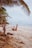 A person is exercising on a sandy beach, using resistance straps attached to a palm tree. The peaceful ocean waves and overcast sky create a serene atmosphere. Palm trees and scattered seaweed line the edge of the beach.