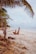 A person is exercising on a sandy beach, using resistance straps attached to a palm tree. The peaceful ocean waves and overcast sky create a serene atmosphere. Palm trees and scattered seaweed line the edge of the beach.