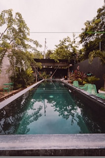 A long, narrow swimming pool is surrounded by lush greenery and outdoor seating. The water reflects the overhanging trees and the sky. There are green lounge chairs and small tables adjacent to the pool, with vibrant plants and a pergola at the end.