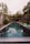 A long, narrow swimming pool is surrounded by lush greenery and outdoor seating. The water reflects the overhanging trees and the sky. There are green lounge chairs and small tables adjacent to the pool, with vibrant plants and a pergola at the end.