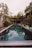 A long, narrow swimming pool is surrounded by lush greenery and outdoor seating. The water reflects the overhanging trees and the sky. There are green lounge chairs and small tables adjacent to the pool, with vibrant plants and a pergola at the end.