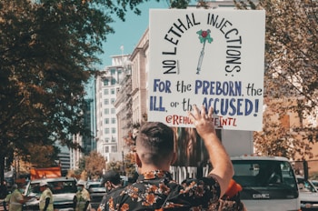 A person is holding a sign in the air at a public demonstration. The sign reads 'No lethal injections: for the preborn, the ill, or the accused!' The scene appears to be on a city street with several buildings, trees, and vehicles in the background. Other people are present, some wearing high-visibility vests.