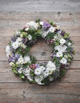 A floral wreath composed of various flowers and greenery sits on a rustic wooden surface. The arrangement includes white daisies, purple hydrangeas, and lush green leaves, creating a natural and earthy feel.