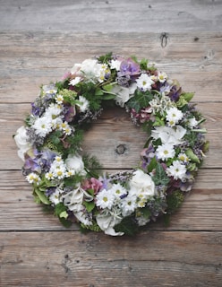 A floral wreath composed of various flowers and greenery sits on a rustic wooden surface. The arrangement includes white daisies, purple hydrangeas, and lush green leaves, creating a natural and earthy feel.