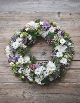A floral wreath composed of various flowers and greenery sits on a rustic wooden surface. The arrangement includes white daisies, purple hydrangeas, and lush green leaves, creating a natural and earthy feel.