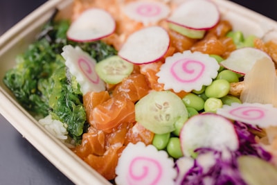 A colorful bento box featuring a mix of fresh ingredients. There are slices of raw salmon, topped with thinly sliced cucumber, radish, and narutomaki with a pink spiral design. Edamame beans, purple cabbage, and green seaweed provide a vibrant contrast, all set on a bed of white rice.