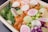 A colorful bento box featuring a mix of fresh ingredients. There are slices of raw salmon, topped with thinly sliced cucumber, radish, and narutomaki with a pink spiral design. Edamame beans, purple cabbage, and green seaweed provide a vibrant contrast, all set on a bed of white rice.