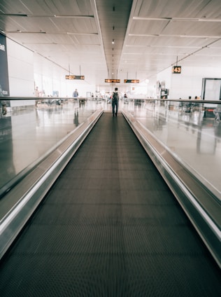 A person walking on an airport moving walkway, with signage overhead indicating gate numbers. The setting is spacious and well-lit with a modern design.