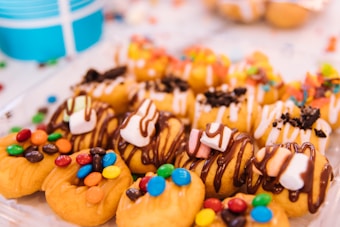 Colorful mini donuts are topped with various candies, marshmallows, and drizzles of chocolate and white icing. The background is soft and blurred, emphasizing the vibrant toppings and textures of the confectionery.