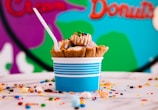 A colorful scene features a blue-striped cup filled with ice cream topped with various toppings, including a waffle cone, whipped cream, candy pieces, and a spoon. The backdrop displays a vibrant mural with shades of green, blue, purple, and orange. The surface around the cup is scattered with different-colored candies and sprinkles.