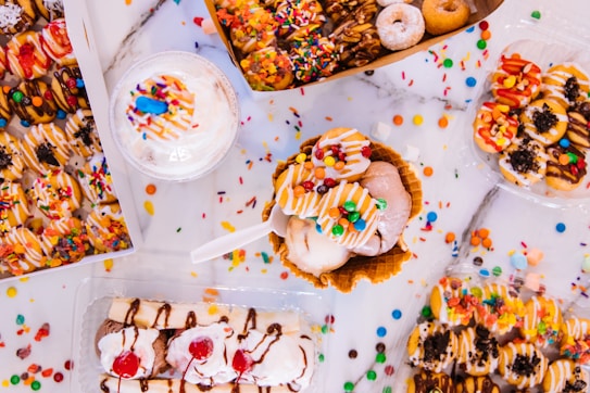 A variety of colorful, decorated donuts and a waffle bowl filled with ice cream are arranged on a table. The donuts are topped with colorful sprinkles, chocolate drizzle, candies, and other toppings. A milkshake with whipped cream and a blue candy sits nearby, while a classic banana split with whipped cream and cherries is also visible.