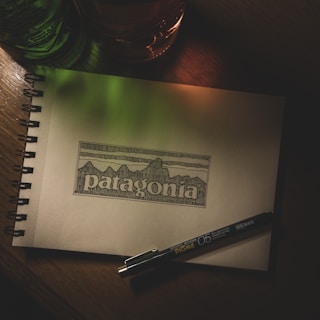 A sketchbook lies open on a wooden surface, displaying an intricate line drawing of mountains with the word 'patagonia' beneath it. Beside the drawing, a Pigma Micron pen is placed, suggesting artistic activity. The scene is dimly lit with green and orange light casting a soft glow, adding depth and atmosphere.