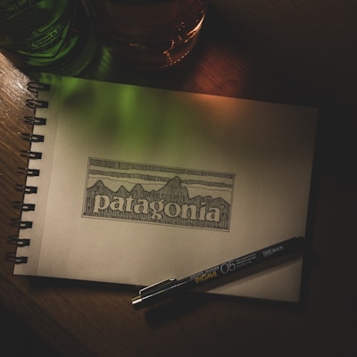 A sketchbook lies open on a wooden surface, displaying an intricate line drawing of mountains with the word 'patagonia' beneath it. Beside the drawing, a Pigma Micron pen is placed, suggesting artistic activity. The scene is dimly lit with green and orange light casting a soft glow, adding depth and atmosphere.