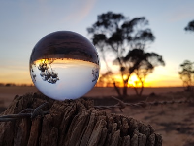 A crystal ball placed on an old piece of wood reflects an inverted image of a sunset and silhouetted trees. The background features a vivid sunset with orange and yellow hues, alongside a tranquil, open landscape. The focus is on the sphere and the play of light within it.