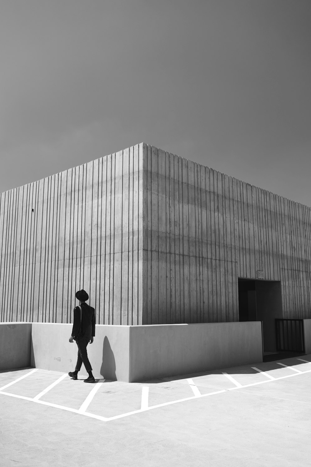 grayscale photo of man in black jacket and pants standing beside building