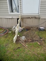 A section of a building exterior is visible, featuring a partially open utility panel revealing wiring and piping, likely for a water heater. Below, there's disturbed earth and scattered debris, including pieces of wood and a metal bowl. Coiled hoses and cables are seen on the grass nearby.