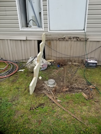 A section of a building exterior is visible, featuring a partially open utility panel revealing wiring and piping, likely for a water heater. Below, there's disturbed earth and scattered debris, including pieces of wood and a metal bowl. Coiled hoses and cables are seen on the grass nearby.