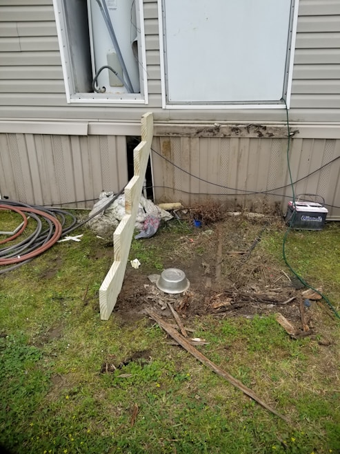 A section of a building exterior is visible, featuring a partially open utility panel revealing wiring and piping, likely for a water heater. Below, there's disturbed earth and scattered debris, including pieces of wood and a metal bowl. Coiled hoses and cables are seen on the grass nearby.