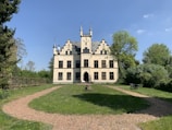 A picturesque, small castle with cream-colored walls features Gothic-style architecture, including intricate spires and arched windows. Surrounding the castle is a lush green lawn, a circular gravel path, and several trees, providing a serene and tranquil setting. The sky is bright blue with a few scattered clouds.