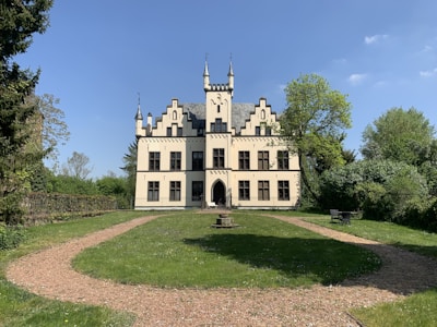 A picturesque, small castle with cream-colored walls features Gothic-style architecture, including intricate spires and arched windows. Surrounding the castle is a lush green lawn, a circular gravel path, and several trees, providing a serene and tranquil setting. The sky is bright blue with a few scattered clouds.